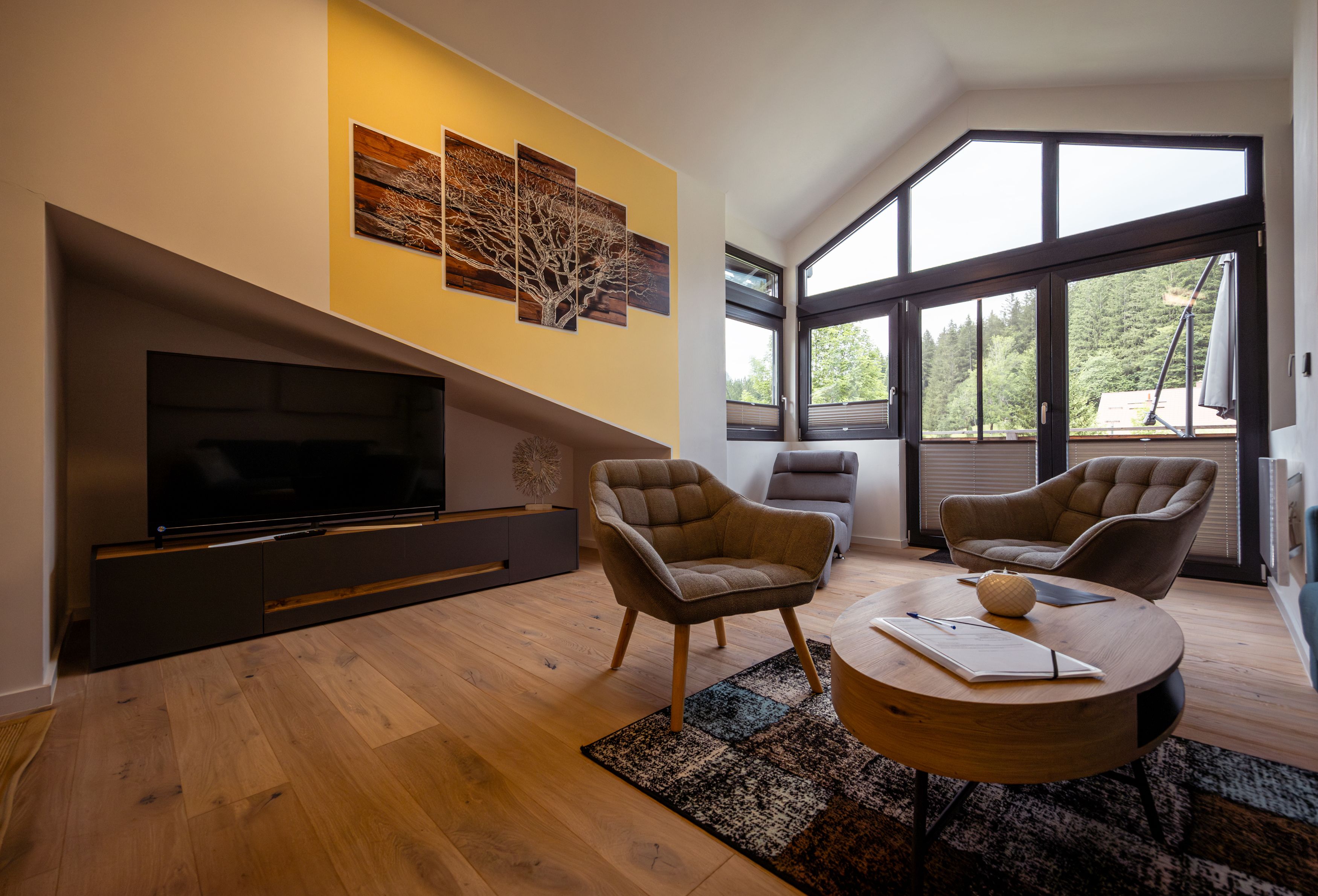 Modernes Wohnzimmer mit großen Fenstern, zwei Sesseln, einem Fernseher und einem runden Couchtisch.