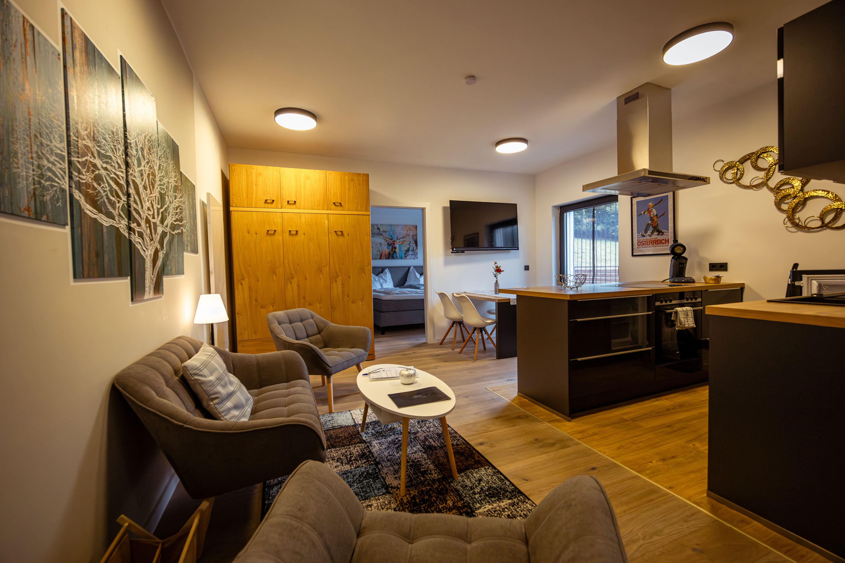 Modern eingerichtetes Wohnzimmer mit Küche, Sofa, Sesseln und Esstisch. Helle Holzmöbel und Wandkunst sind zu sehen.