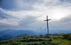 A majestic summit cross towers over the rolling hills, surrounded by a breathtaking mountain landscape. The clouds gently drift across the sky and lend the scenery a peaceful atmosphere, while the fresh mountain air invites you to go hiking.