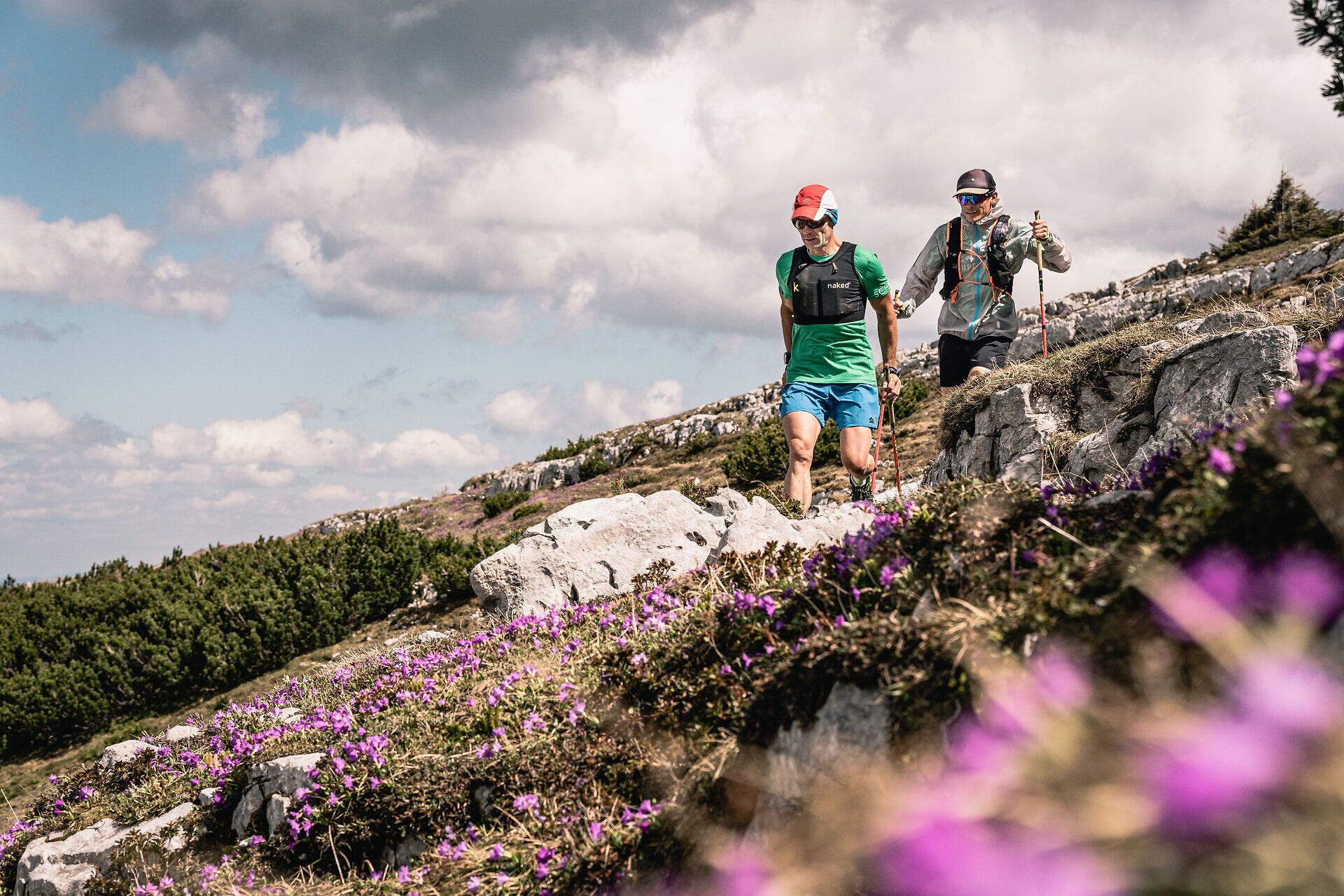 Zwei Trailrunner durchqueren die blühenden Wiesen und felsigen Pfade, während die majestätischen Berge im Hintergrund aufragen. Die frische Bergluft und die bunten Wildblumen schaffen eine einladende Atmosphäre für Abenteuerlustige und Naturliebhaber. Hier wird der Sommer in den Bergen zum unvergesslichen Erlebnis.