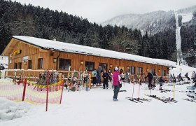 Ski hut in the snow with skiers and equipment in front of it.